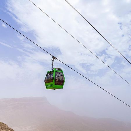 Tolip Resort El Galala Majestic Айн Сохна Екстериор снимка The photo shows a green cable car suspended in the air, traveling along overhead cables. The backdrop features a hazy landscape, likely mountainous or elevated terrain, with a cloudy sky. The scene suggests a scenic transportation method, possibly us