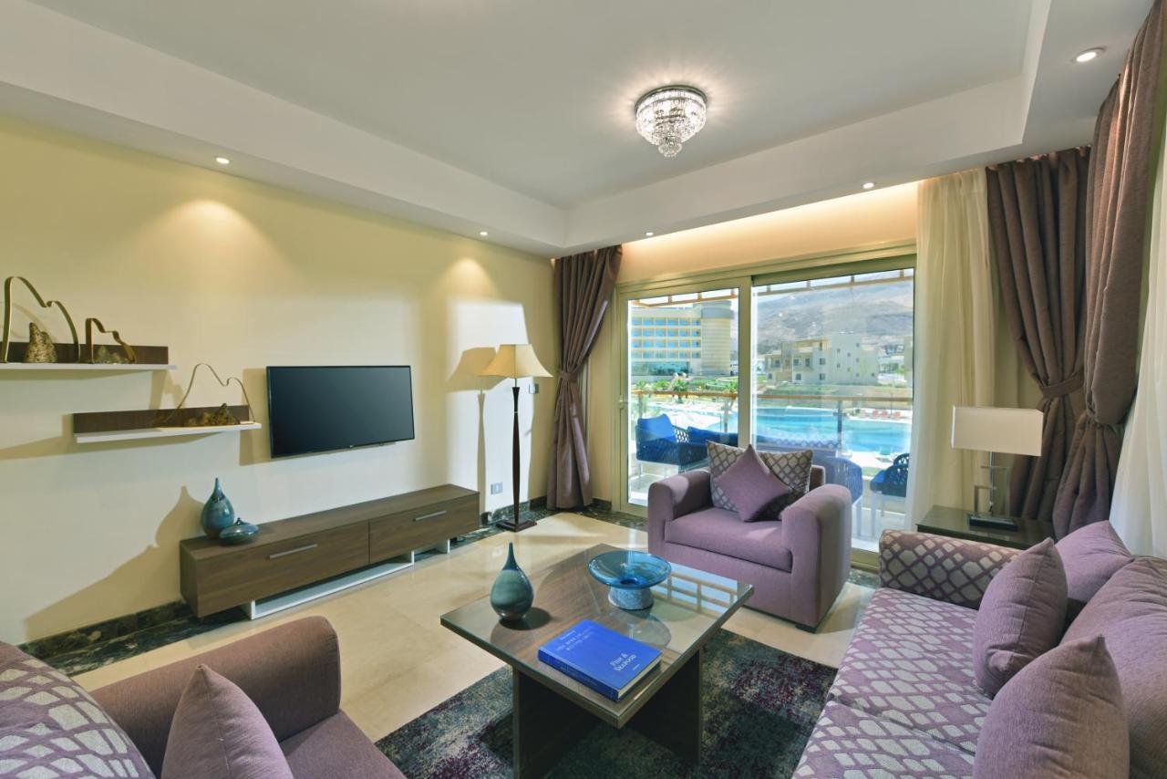Tolip Resort El Galala Majestic Айн Сохна Екстериор снимка The photo shows a modern living room that features a stylish and cozy design. There are two purple sofas with decorative cushions arranged around a coffee table. The coffee table has a blue book and a decorative item on it. A flat-screen TV is mounte