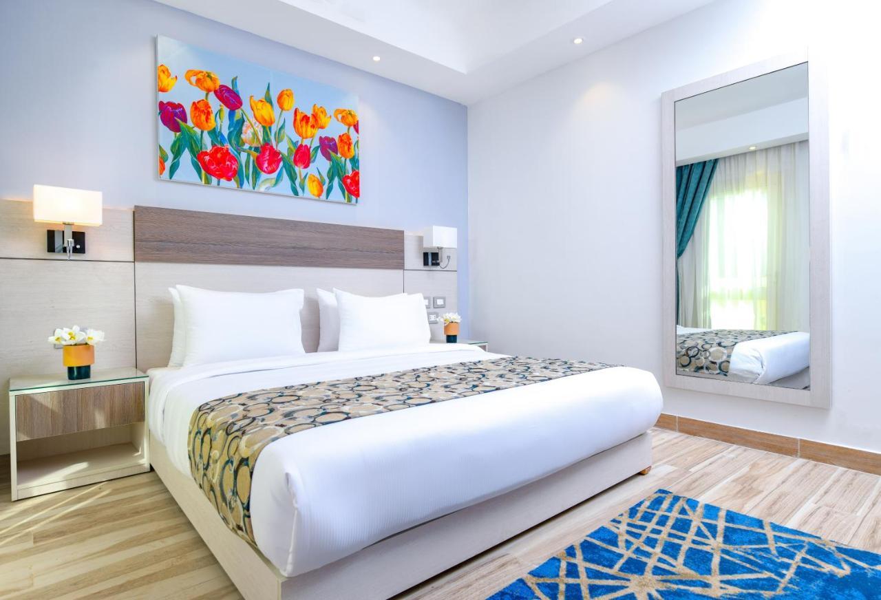 Tolip Resort El Galala Majestic Айн Сохна Екстериор снимка The photo shows a modern hotel bedroom. It features a large bed with white linens and a decorative throw blanket. The room has light wooden flooring and is bright, with ample natural light coming from a window. There is a colorful painting of flowers