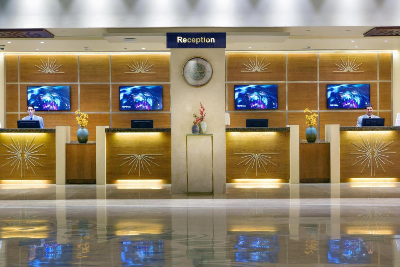 Tolip Resort El Galala Majestic Айн Сохна Екстериор снимка The image shows a hotel reception area. There are several reception desks lined up against a wall with decorative paneling. Each desk has a staff member, and above the desks, there are screens displaying information. A clock is visible on the wall, a
