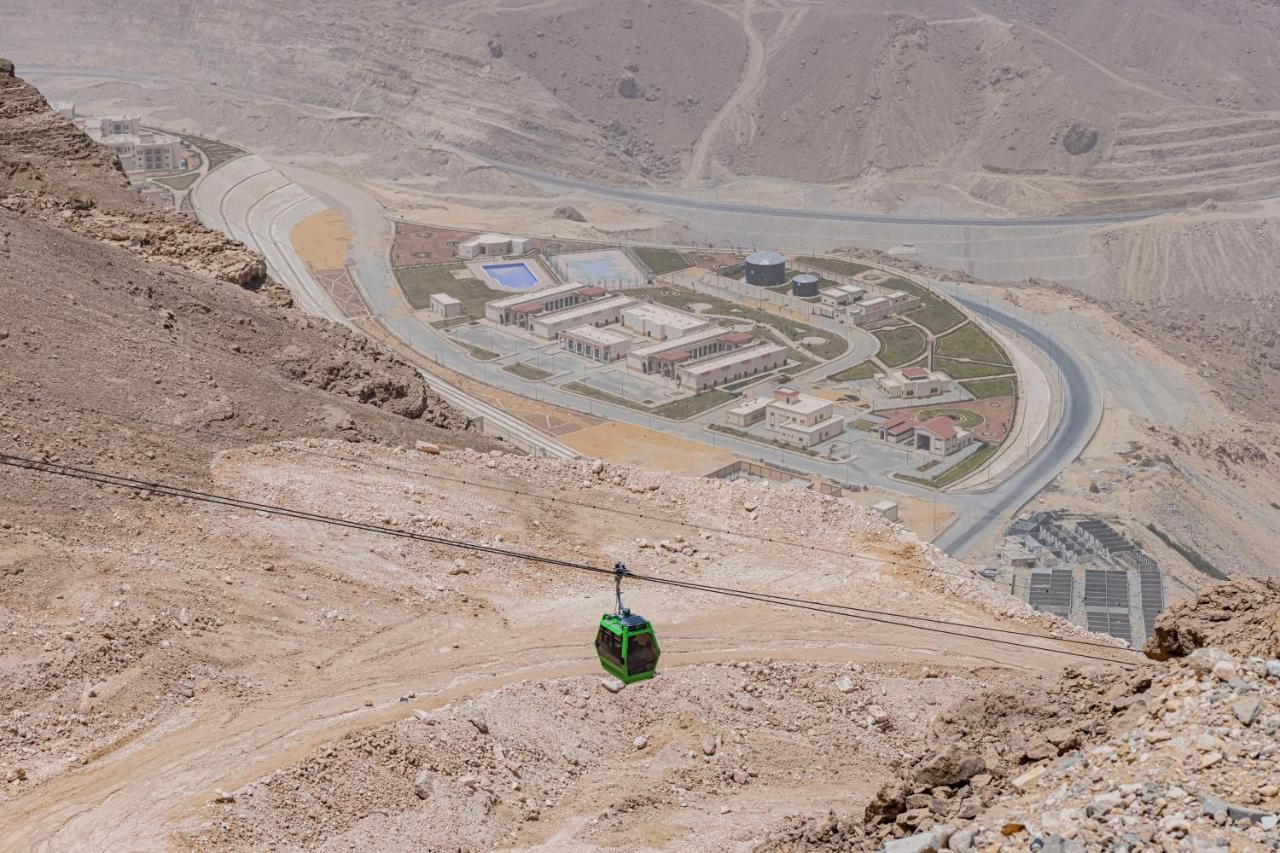 Tolip Resort El Galala Majestic Айн Сохна Екстериор снимка The photo shows a mountainous landscape with a steep, dry terrain. In the foreground, there is a green cable car or gondola traveling along a cable. Below, a complex of buildings and roads can be seen, likely part of a resort or recreational area. Th