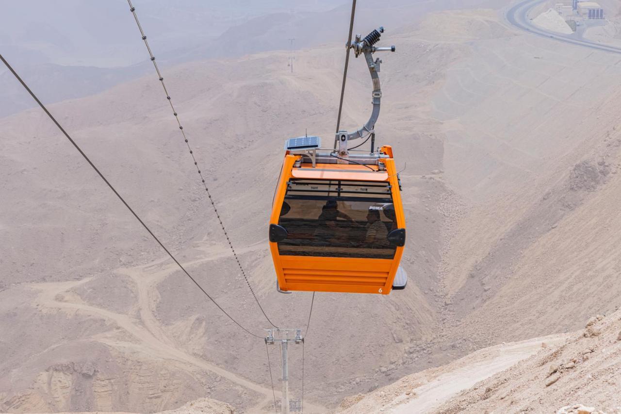 Tolip Resort El Galala Majestic Айн Сохна Екстериор снимка The photo shows an orange cable car suspended in the air, traveling along a set of cables. It appears to be located in a mountainous or hilly area, with rugged terrain and dry, rocky landscapes visible below. The background features slopes that seem 
