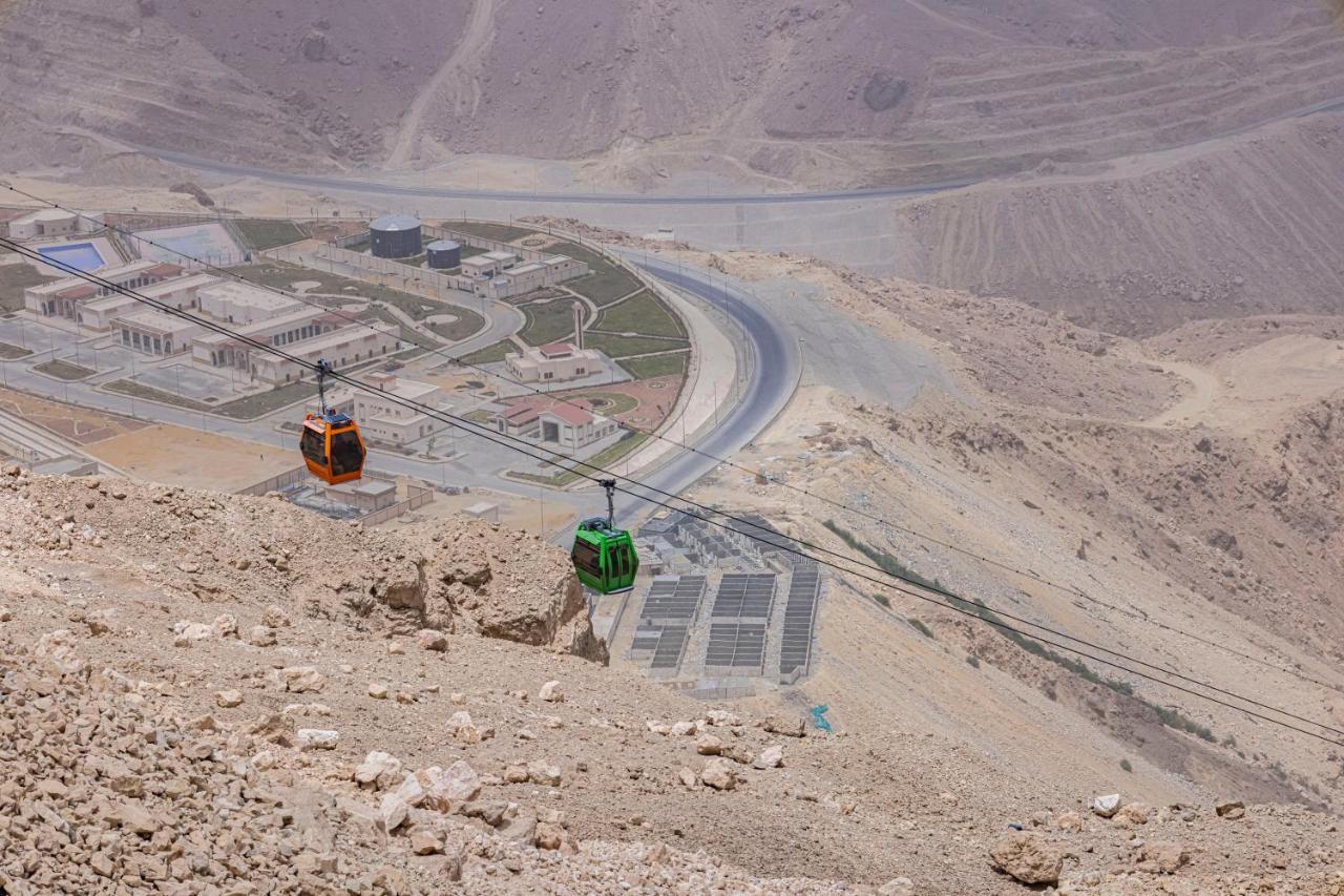 Tolip Resort El Galala Majestic Айн Сохна Екстериор снимка The photo shows a scenic view from a high elevation, featuring a cable car system. Two cable cars are visible, one orange and one green, traveling along cables down the mountainside. Below, there appears to be a developed area with roads and light st
