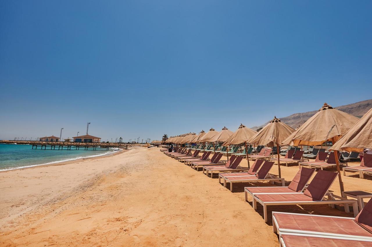 Tolip Resort El Galala Majestic Айн Сохна Екстериор снимка The photo shows a scenic beach with a stretch of golden sand under a clear blue sky. There are several beach loungers arranged in rows, each accompanied by thatched umbrellas providing shade. In the background, there is a wooden pier extending into t