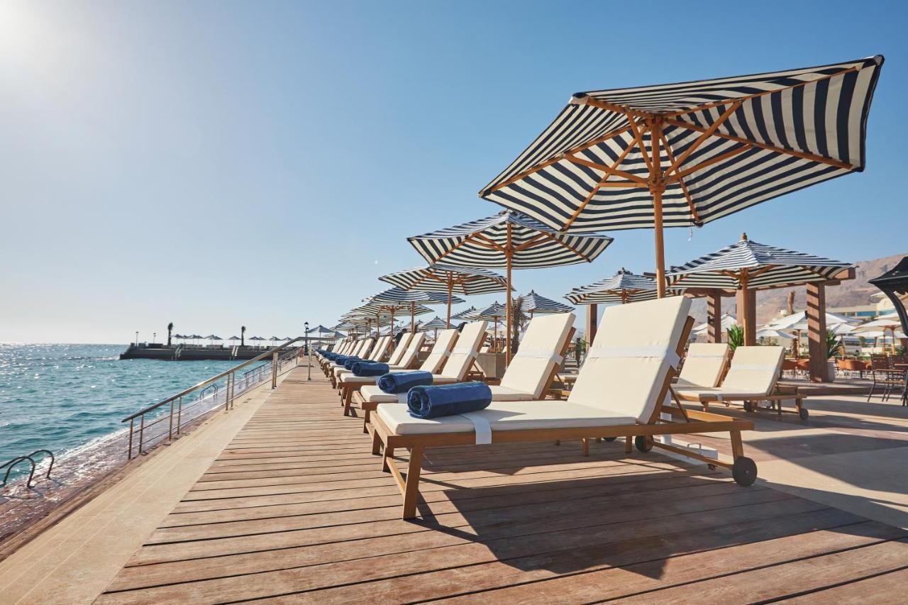 Tolip Resort El Galala Majestic Айн Сохна Екстериор снимка The photo depicts a serene beachside setting featuring a row of sun loungers arranged on a wooden deck. Each lounger is equipped with a folded towel and shaded by striped umbrellas. In the background, the water sparkles under bright sunlight, and the