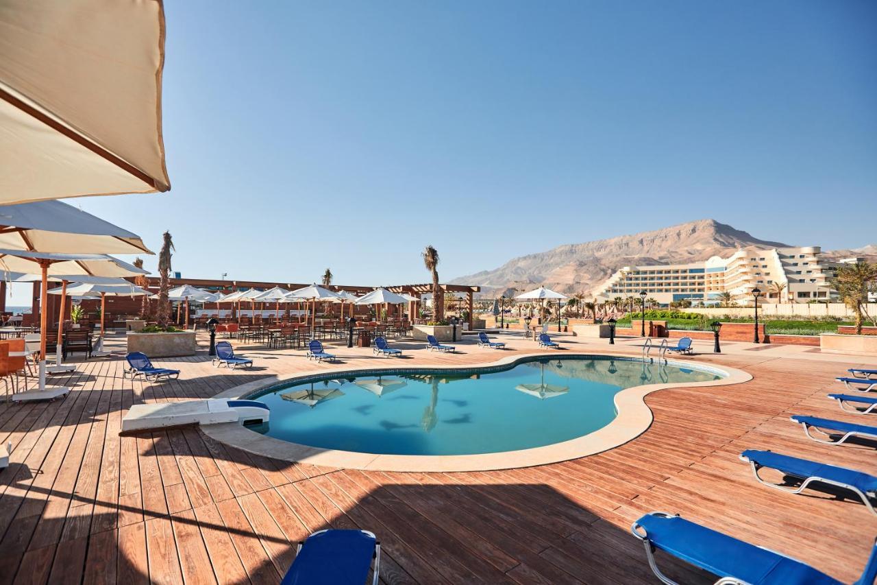 Tolip Resort El Galala Majestic Айн Сохна Екстериор снимка The photo shows a swimming pool area surrounded by sun loungers and umbrellas. The pool has a unique, curvy shape and is situated on a wooden deck. In the background, there are several buildings, likely part of a resort or hotel, and a mountainous la
