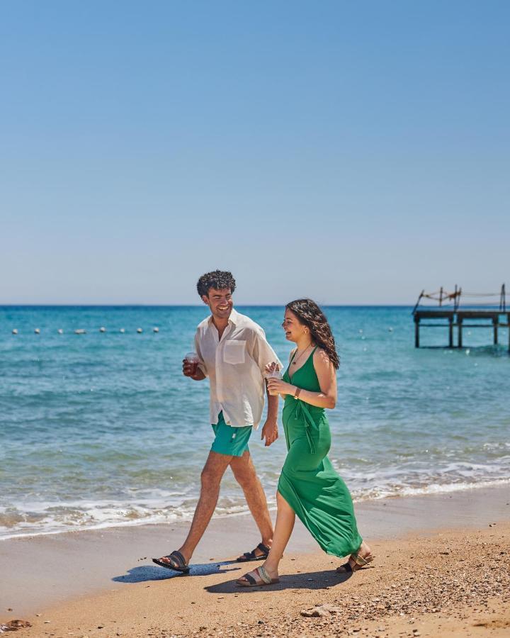 Tolip Resort El Galala Majestic Айн Сохна Екстериор снимка The photo shows a couple walking along a beach. The man is wearing a white short-sleeved shirt and green shorts, while the woman is dressed in a flowing green dress. They both appear to be enjoying the pleasant weather, with the ocean in the backgrou