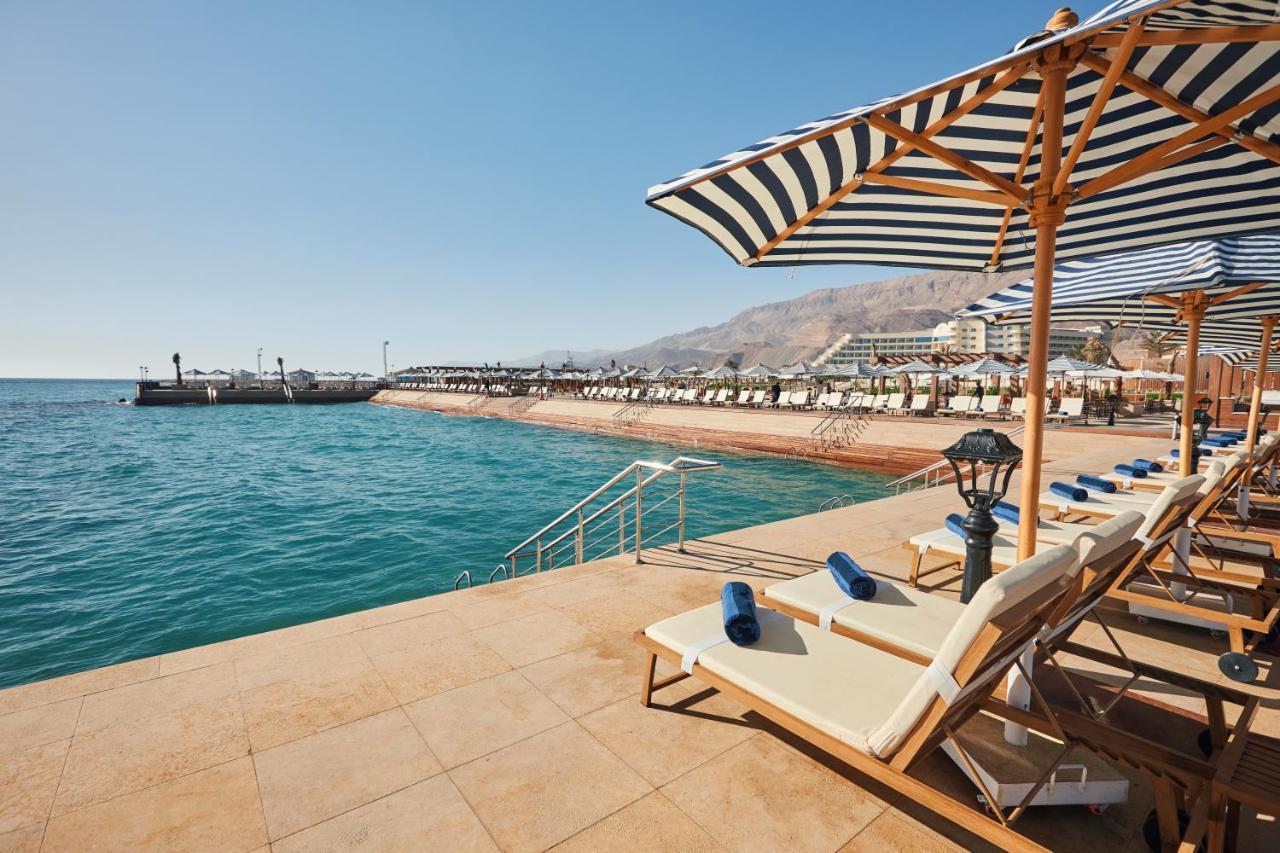 Tolip Resort El Galala Majestic Айн Сохна Екстериор снимка The photo shows a scenic beachside area with clear blue water. In the foreground, there are several sun loungers set up on a tiled surface, with blue towels neatly rolled on each one. The loungers are shaded by striped umbrellas. In the distance, a w