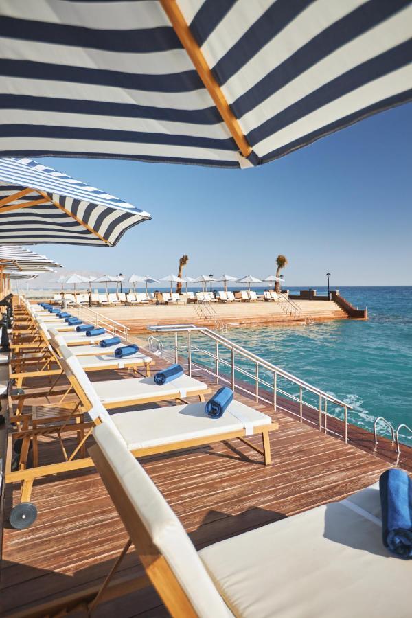 Tolip Resort El Galala Majestic Айн Сохна Екстериор снимка The photo depicts a serene outdoor area by the waterfront. There are several lounge chairs arranged in a row, each equipped with blue towels. Large, striped umbrellas provide shade over some of the chairs. In the background, there is a wooden deck th