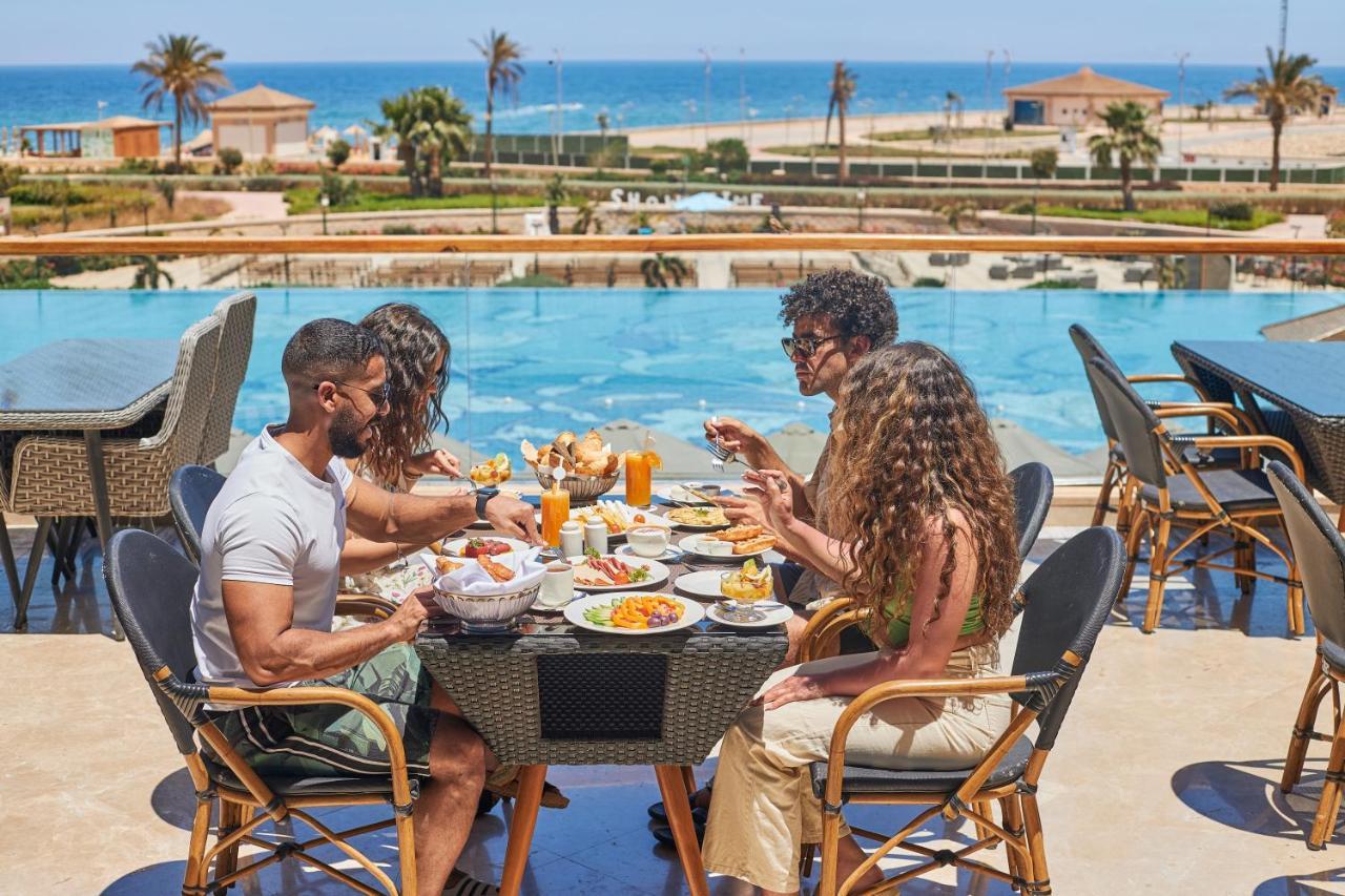 Tolip Resort El Galala Majestic Айн Сохна Екстериор снимка The photo shows a group of five people enjoying a meal together outdoors, likely at a restaurant or cafe. They are seated at a table with a beautiful view of a swimming pool and the sea in the background. The table is filled with various dishes, and 