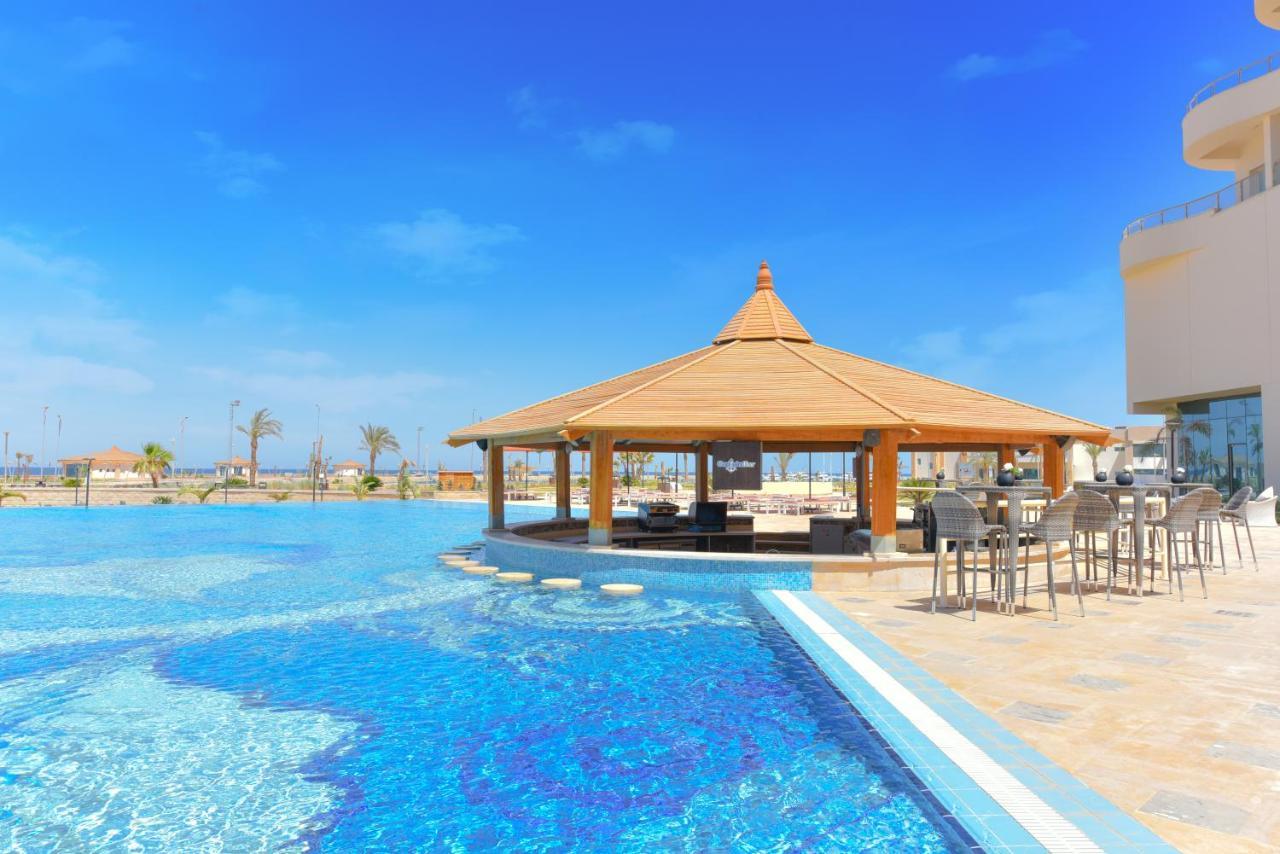 Tolip Resort El Galala Majestic Айн Сохна Екстериор снимка The photo shows a beautiful outdoor setting featuring a large swimming pool with clear blue water. In the center, there is a thatched-roof gazebo, which serves as a shaded seating area. Surrounding the gazebo are lounge chairs and a bar area with hig