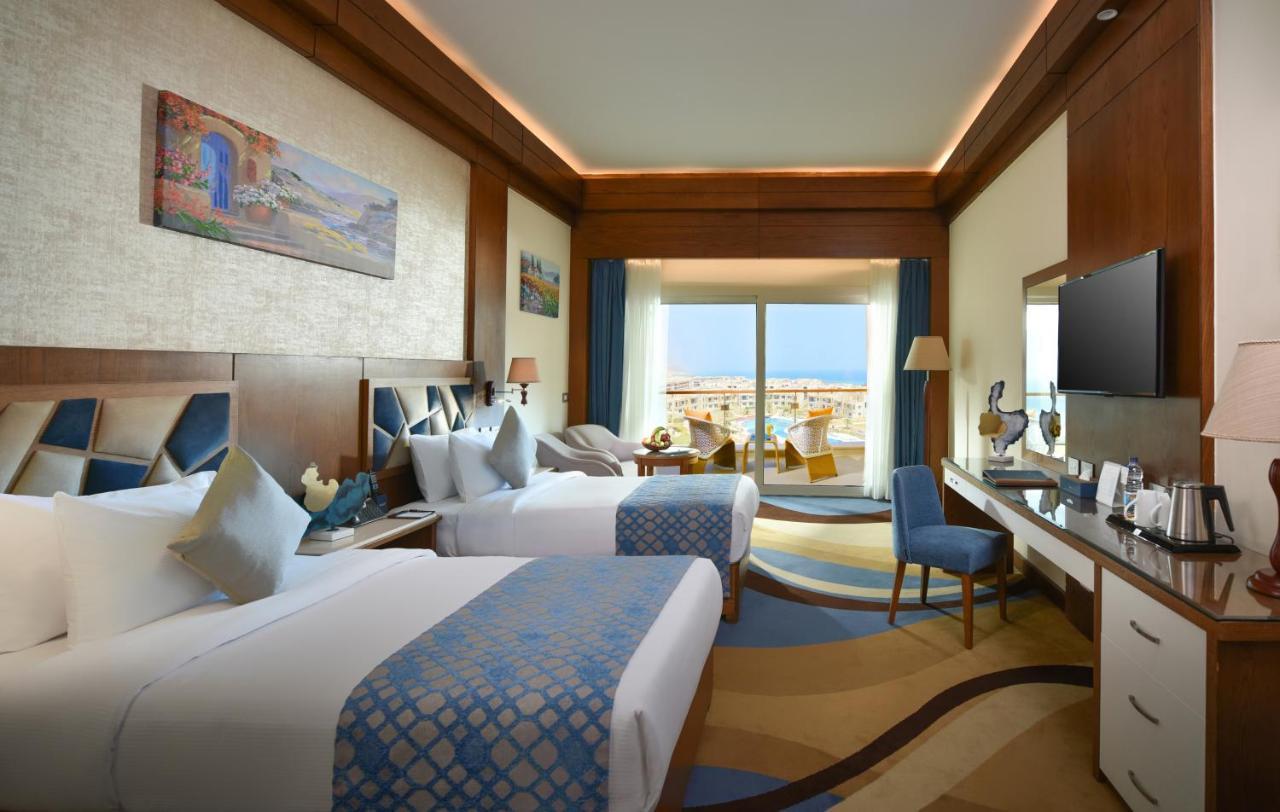 Tolip Resort El Galala Majestic Айн Сохна Екстериор снимка The photo shows a well-decorated hotel room featuring two beds with decorative pillows. The room has wooden accents and modern furnishings, including a desk, a television, and lamps. Large windows flood the space with natural light, offering a view o