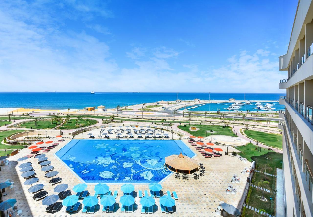Tolip Resort El Galala Majestic Айн Сохна Екстериор снимка The photo shows a beautiful outdoor view of a resort area featuring a large swimming pool with a decorative underwater design. Surrounding the pool are numerous sun loungers and umbrellas in a variety of colors. In the background, there is a scenic c