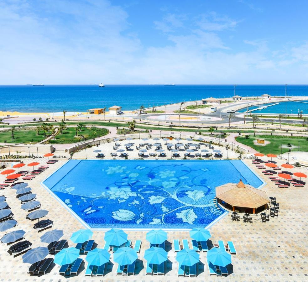 Tolip Resort El Galala Majestic Айн Сохна Екстериор снимка The photo features a large swimming pool with a decorative design on its surface, surrounded by sun loungers and umbrellas. In the background, there is a view of the sea, with a clear blue sky and a few scattered clouds. The area is well-maintained, 