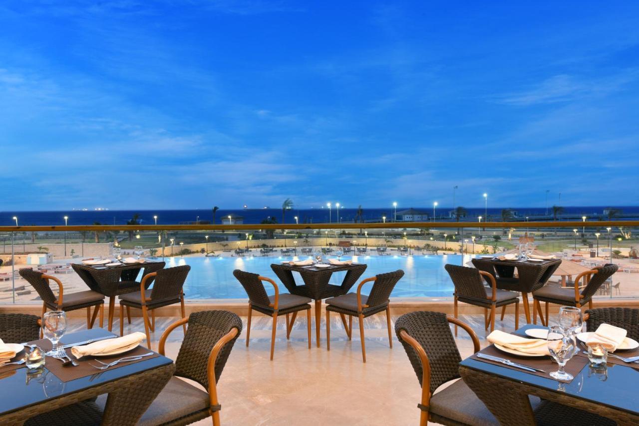 Tolip Resort El Galala Majestic Айн Сохна Екстериор снимка The photo shows an outdoor dining area with several neatly arranged tables and chairs. The tables are elegantly set with cutlery and plates, suggesting a fine dining experience. In the background, there is a large swimming pool that reflects the blue
