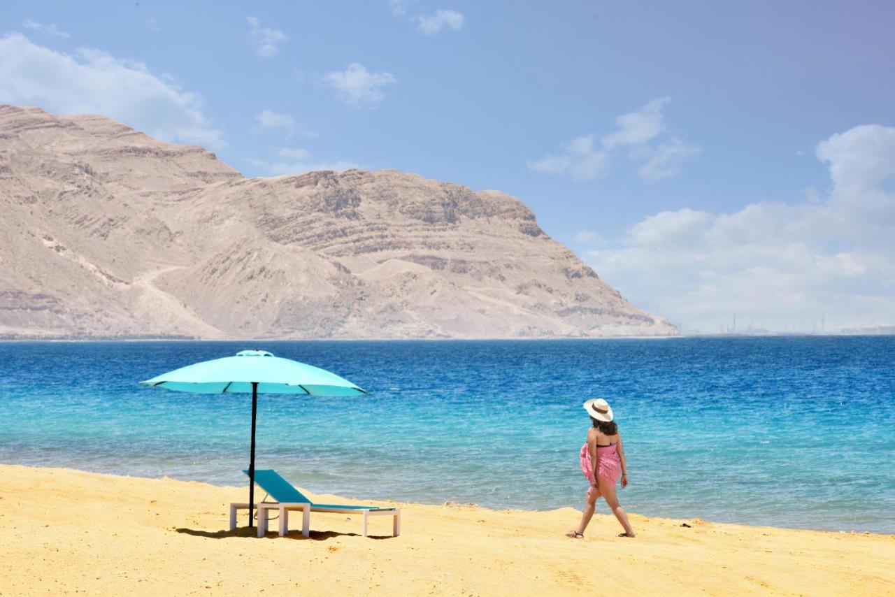 Tolip Resort El Galala Majestic Айн Сохна Екстериор снимка The photo depicts a serene beach scene. In the foreground, there is a sandy shore with a single beach chair and an umbrella in a light blue color. A person dressed in a pink outfit is walking along the shoreline, enjoying the beach atmosphere. In the